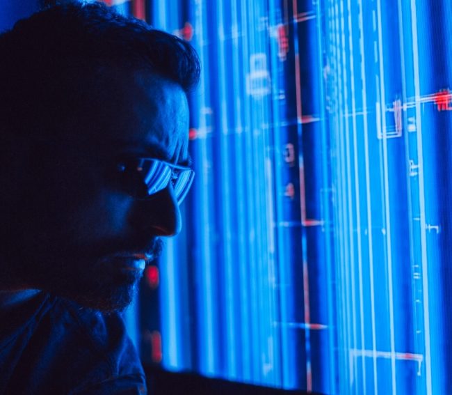 Hombre con lentes de tecnología observando pantallas de servidores o código con luces azules