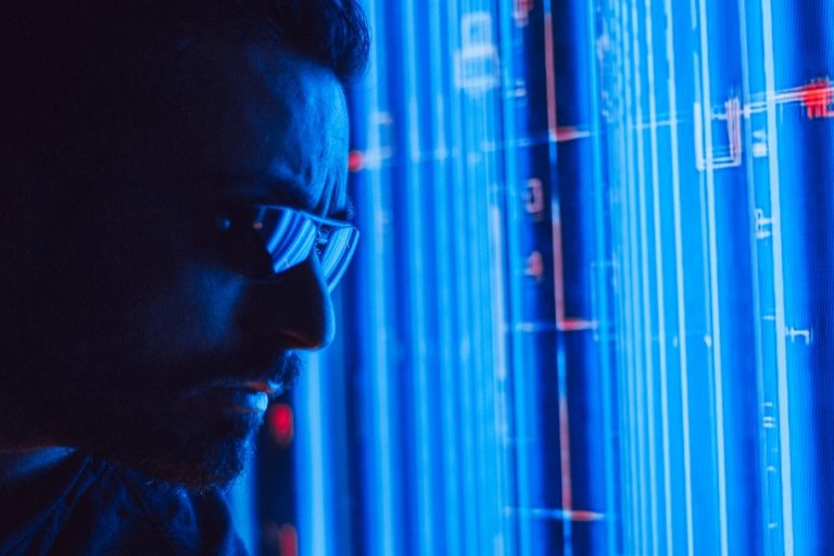 Hombre con lentes de tecnología observando pantallas de servidores o código con luces azules